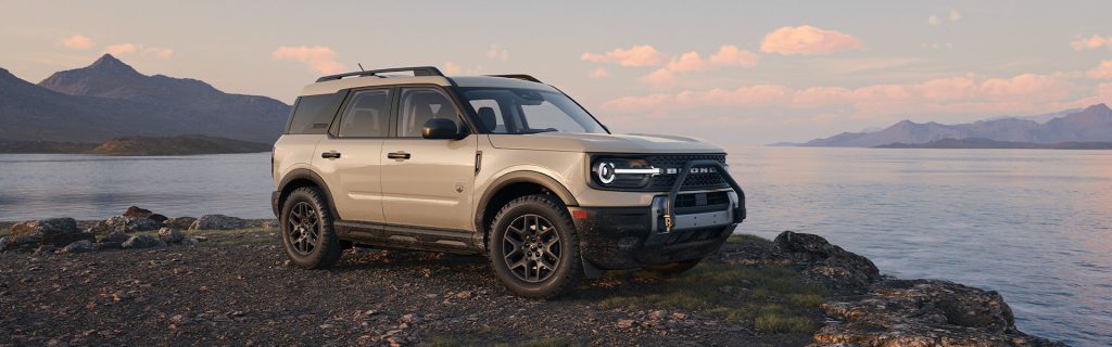 2025 Ford Bronco Sport parked on a rocky lakeside terrain during sunset, showcasing its rugged design, beige exterior, and off-road stance with mountains and calm water in the background.