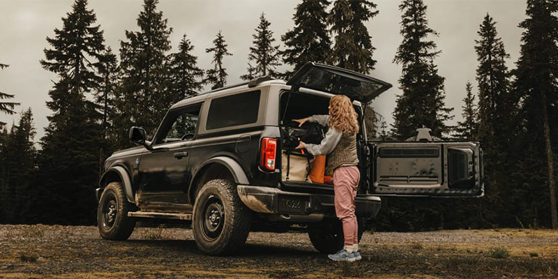 A woman with curly hair packing supplies into the back of a black 2025 Ford Bronco in a forest.